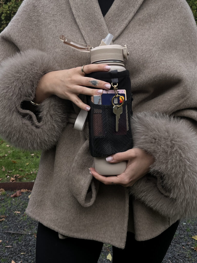 Person holding a reusable water bottle fitted with the Alera bottle sleeve, showing the mesh pocket and key clip designed for secure, on-the-go storage.