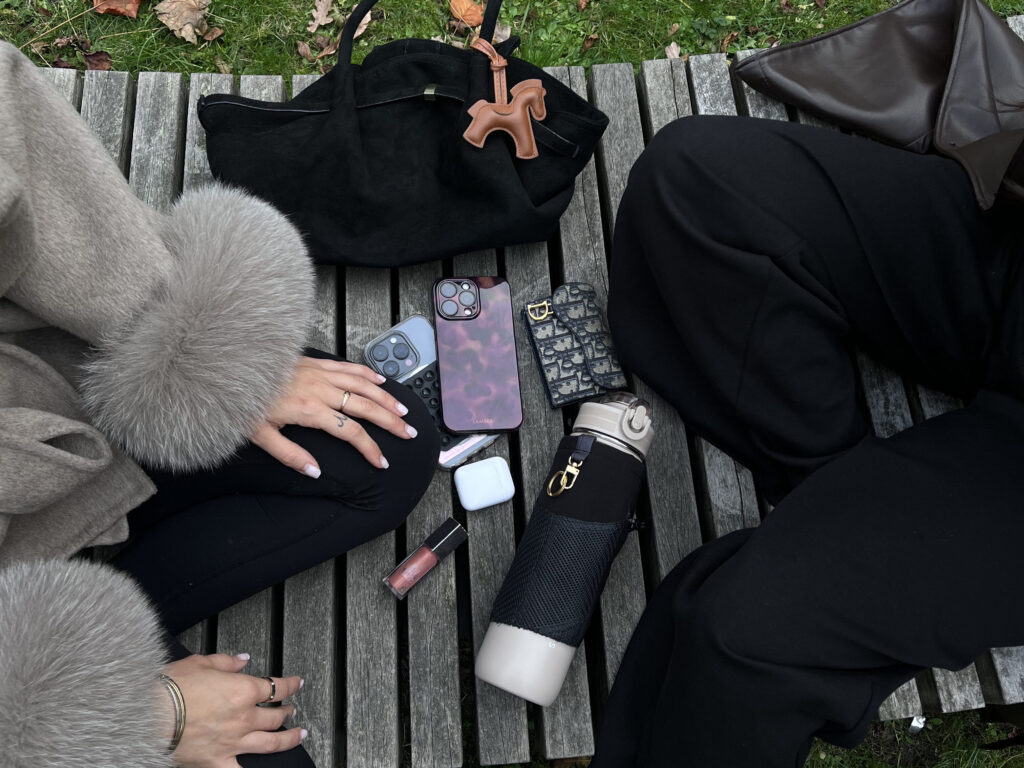 Items spread out on a bench beside friends, with the Alera bottle sleeve holding essentials like keys and small accessories, showing its practical on-the-go storage.